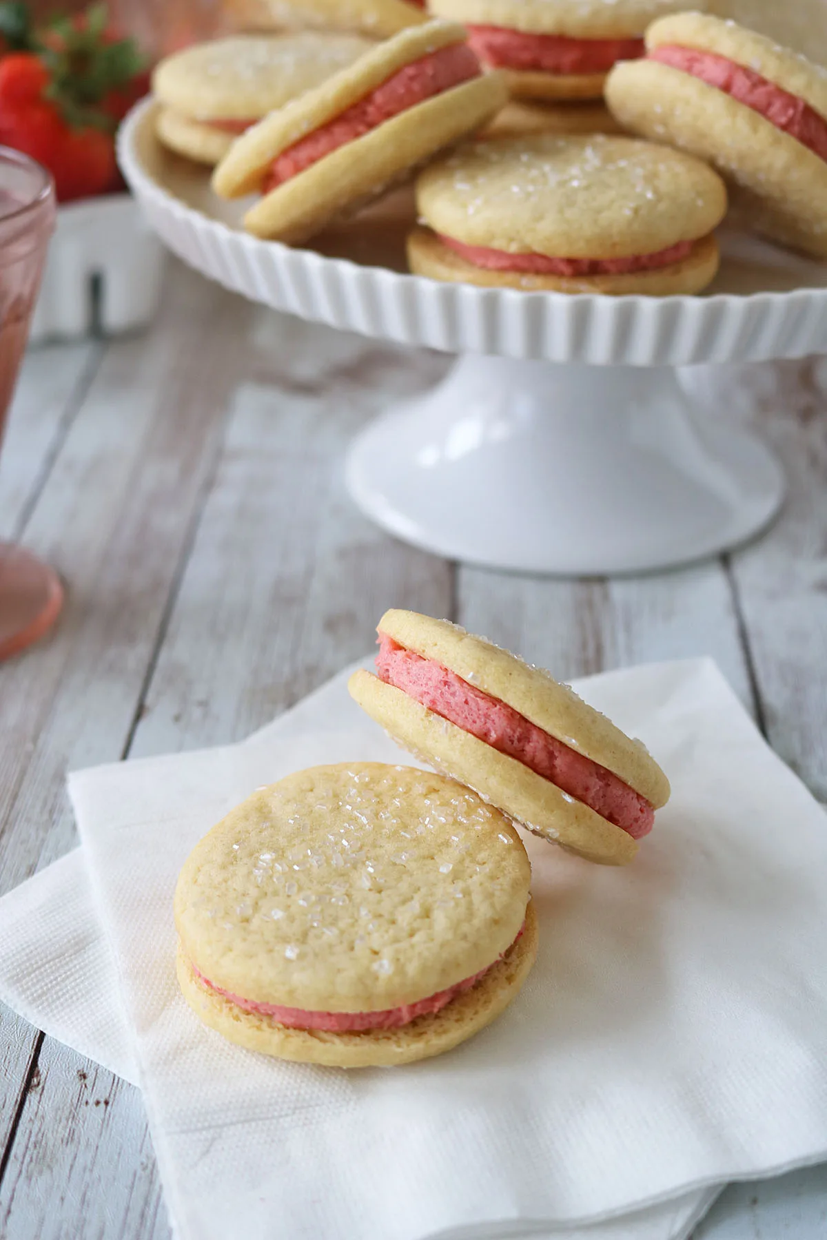 Two Strawberry Shortcake Cookies with pink filling rest on white napkins, while more of these sparkling sugar-topped cookies are displayed on a white cake stand in the background.