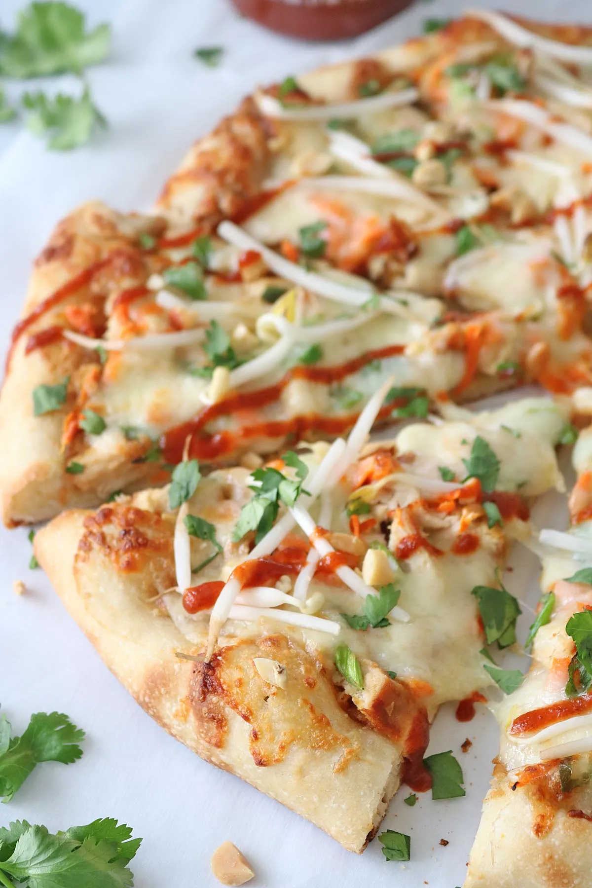 A close-up of sliced pizza topped with melted cheese, bean sprouts, chopped cilantro, peanuts, and a drizzle of red sauce, on a white surface with scattered cilantro leaves.