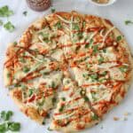 A round pizza topped with cheese, chopped cilantro, bean sprouts, peanuts, and a drizzle of sriracha sauce, with extra peanuts in a bowl and cilantro leaves scattered around on white parchment paper.
