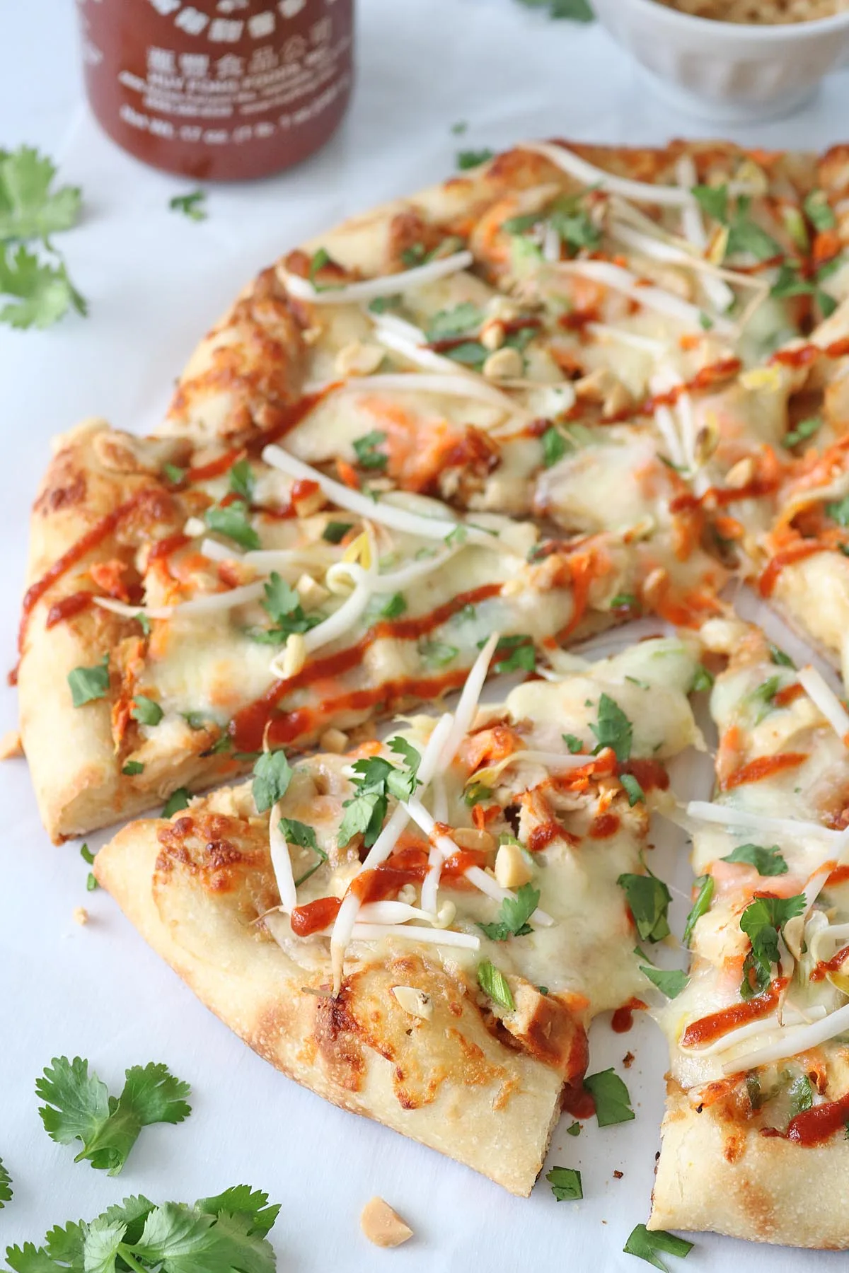 A close-up of a sliced pizza topped with melted cheese, cilantro, peanuts, bean sprouts, and drizzled with sriracha sauce. Fresh cilantro leaves and a sriracha bottle are visible in the background.