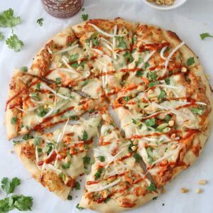 A round pizza topped with cheese, chopped cilantro, bean sprouts, peanuts, and a drizzle of sriracha sauce, with extra peanuts in a bowl and cilantro leaves scattered around on white parchment paper.