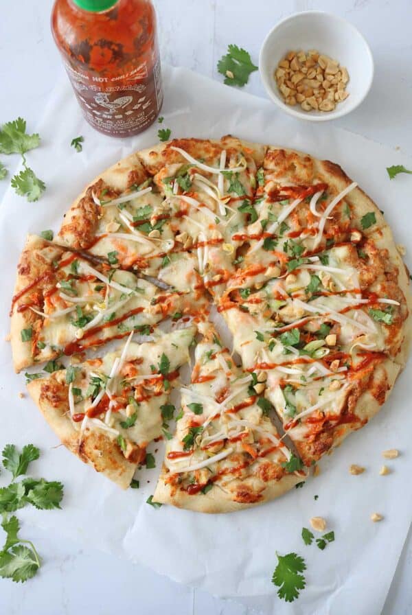 A round pizza topped with cheese, chopped cilantro, bean sprouts, peanuts, and a drizzle of sriracha sauce, with extra peanuts in a bowl and cilantro leaves scattered around on white parchment paper.