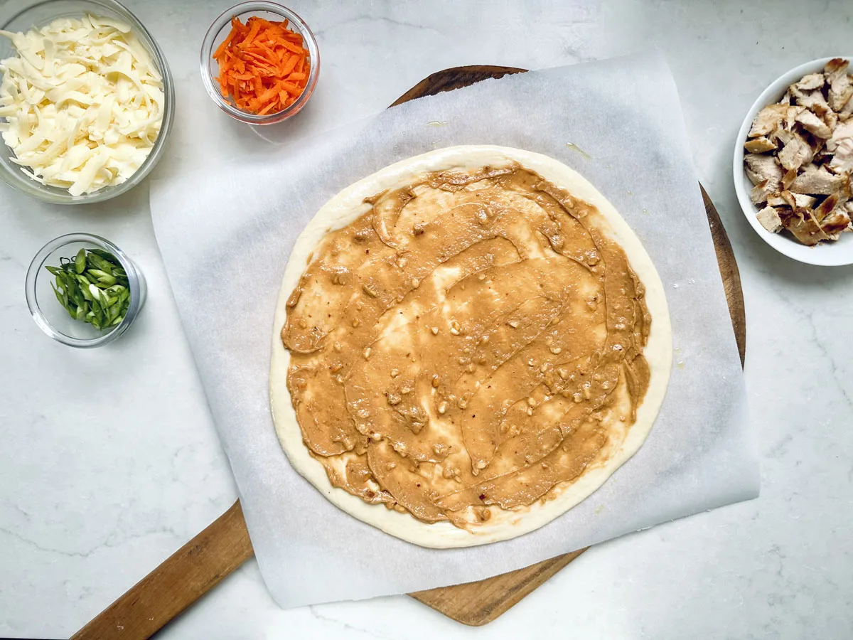 A pizza crust on parchment paper is spread with peanut sauce. Surrounding the crust are bowls containing shredded cheese, sliced green onions, shredded carrots, and chopped cooked chicken. All items are on a white surface.