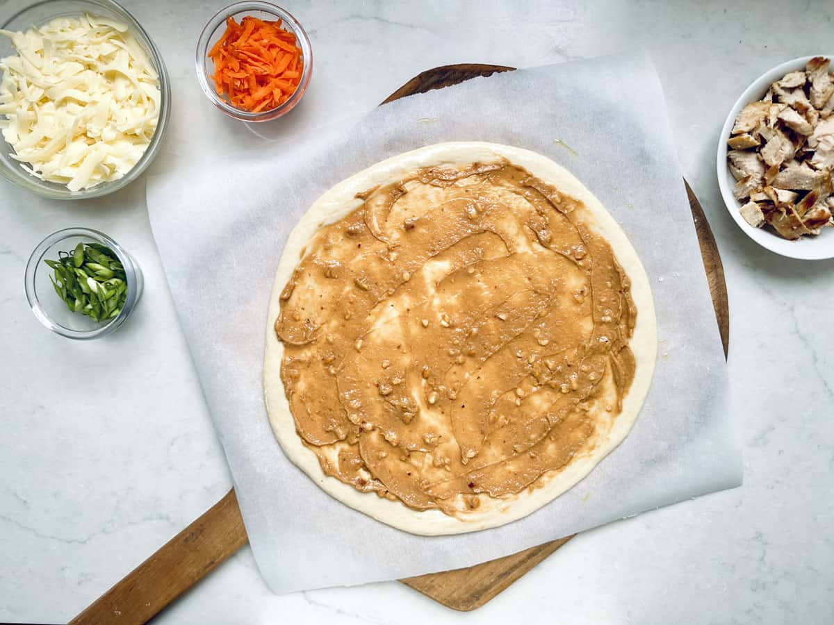 A pizza crust on parchment paper is spread with peanut sauce. Surrounding the crust are bowls containing shredded cheese, sliced green onions, shredded carrots, and chopped cooked chicken. All items are on a white surface.