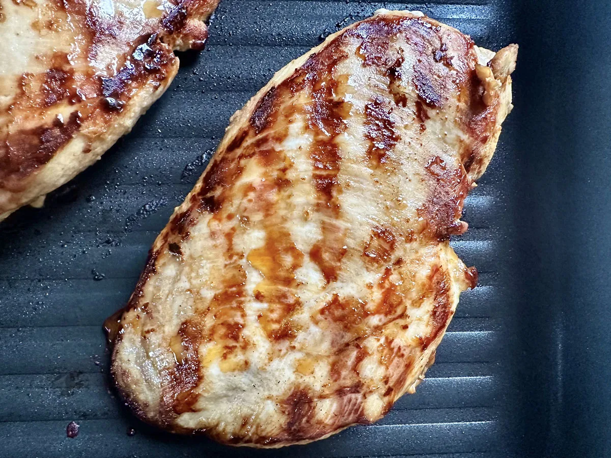 A grilled chicken breast with visible char marks sits on a dark ridged grill pan. The meat appears cooked and juicy with a golden-brown, slightly glossy surface.
