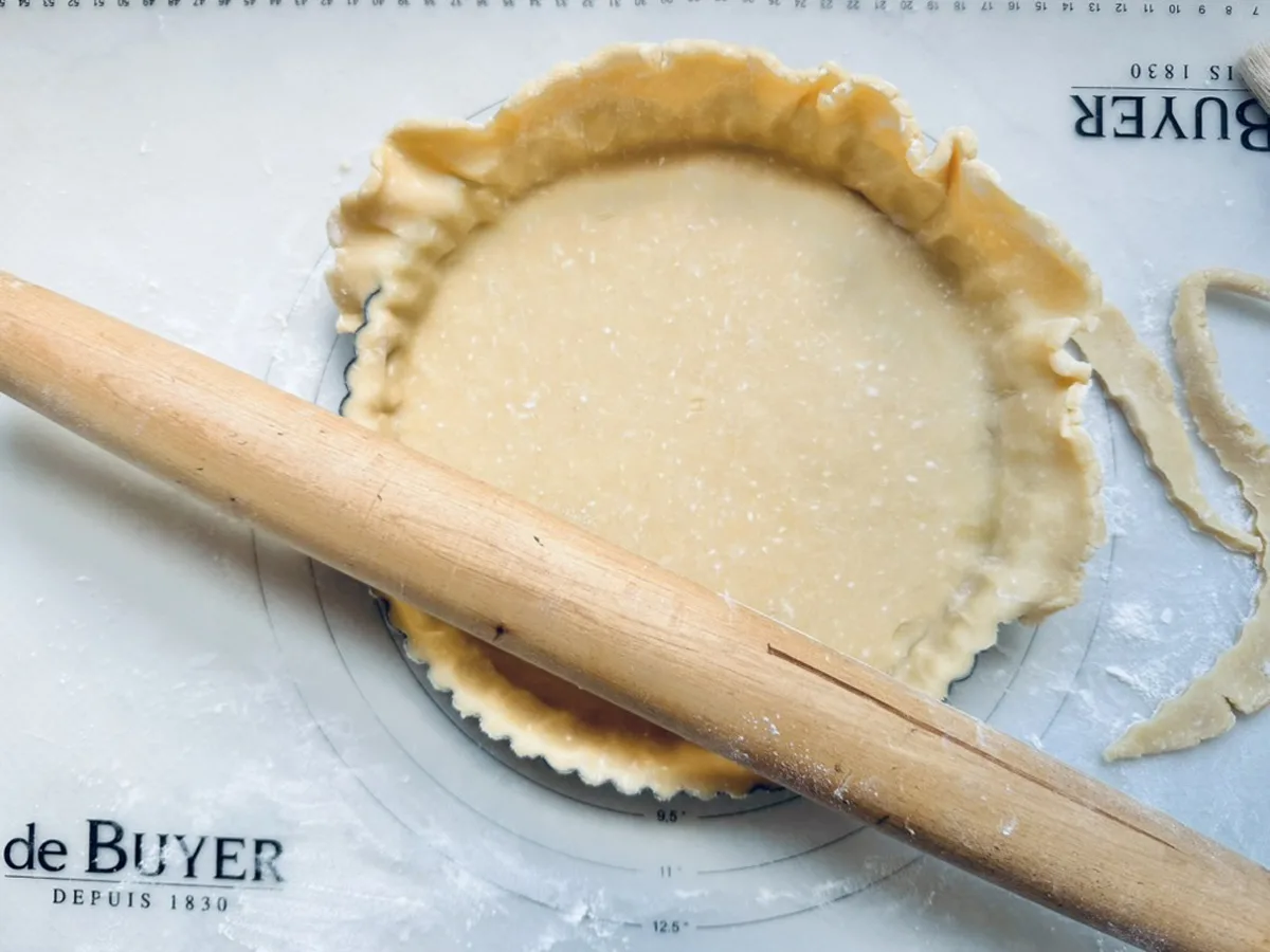 A rolling pin rests on a floured pastry mat next to an unbaked pie crust in a round dish, with excess dough trimmed around the edges.