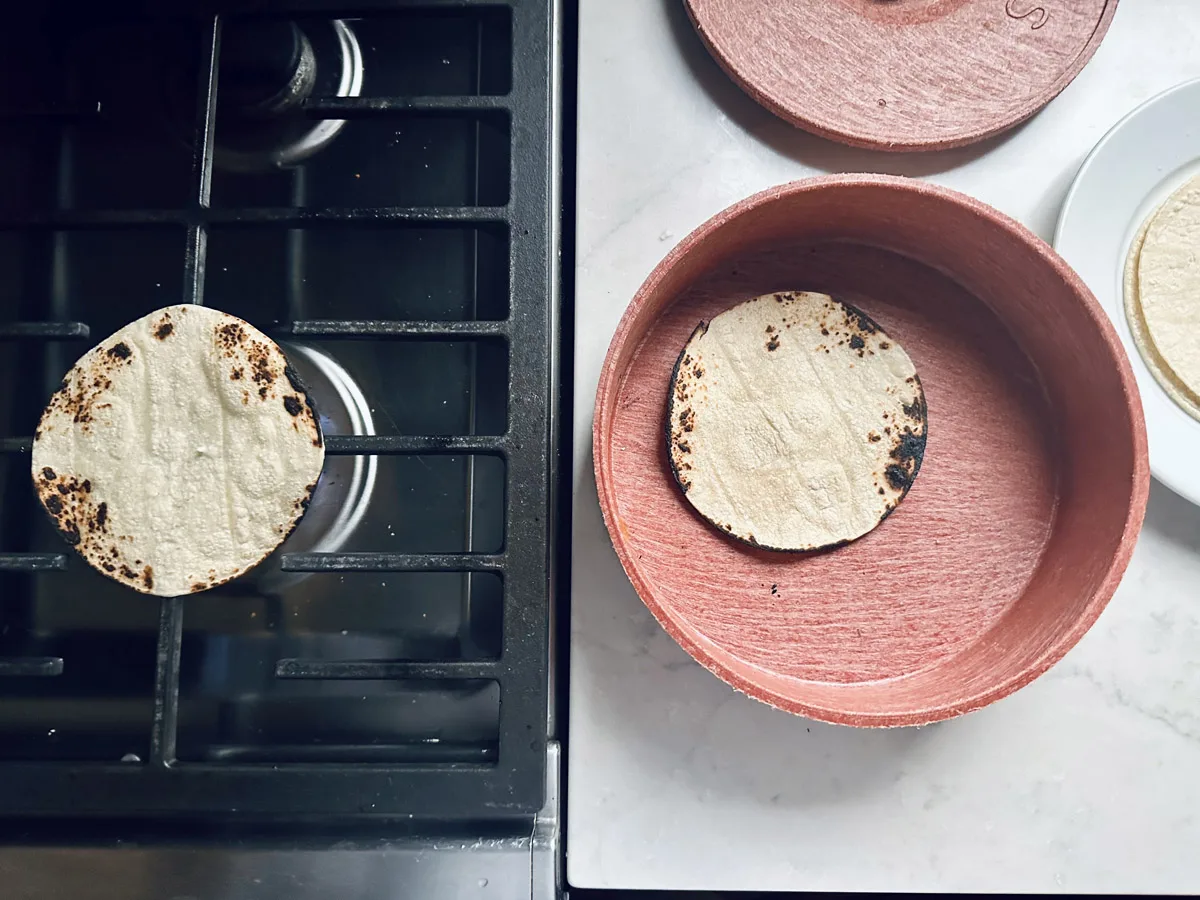 A corn tortilla is warming on a gas stove burner while another tortilla sits in a red container on a countertop nearby. A plate with more tortillas is partially visible to the right.