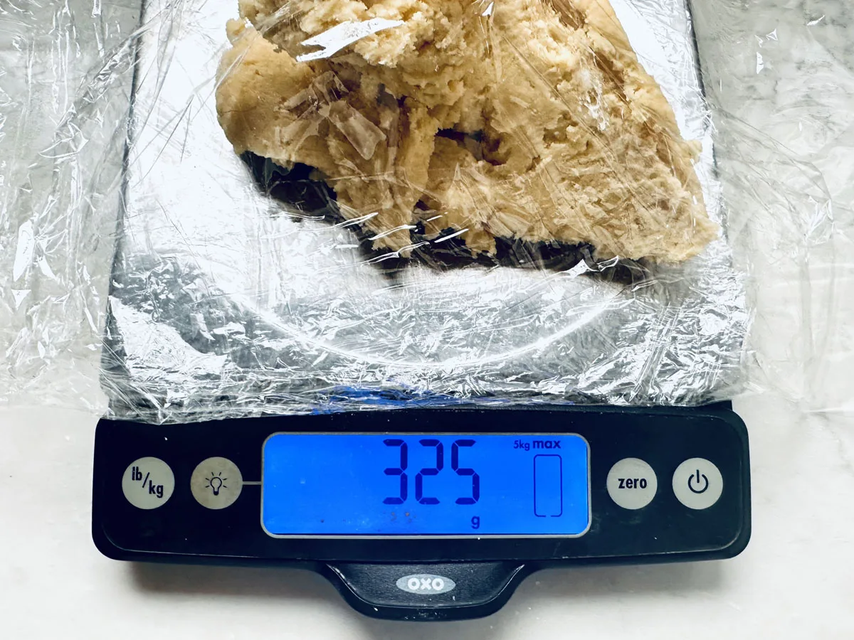 A digital kitchen scale displays 325 grams, weighing dough for Strawberry Shortcake Cookies on a plastic-wrapped surface. The black scale features buttons and a blue-lit screen, perfect for precise baking.