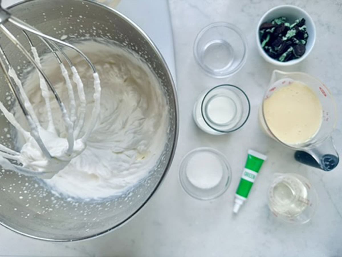 A metal mixing bowl with whipped cream and a whisk is surrounded by small bowls of ingredients, including sugar, milk, chocolate pieces, a tube of green food coloring, and a measuring cup with a light yellow liquid.