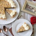 A slice of chocolate chip cookie pie with whipped cream on a white plate, alongside the rest of the pie, two spoons, red roses, and a framed vintage photo on a rustic wooden table.