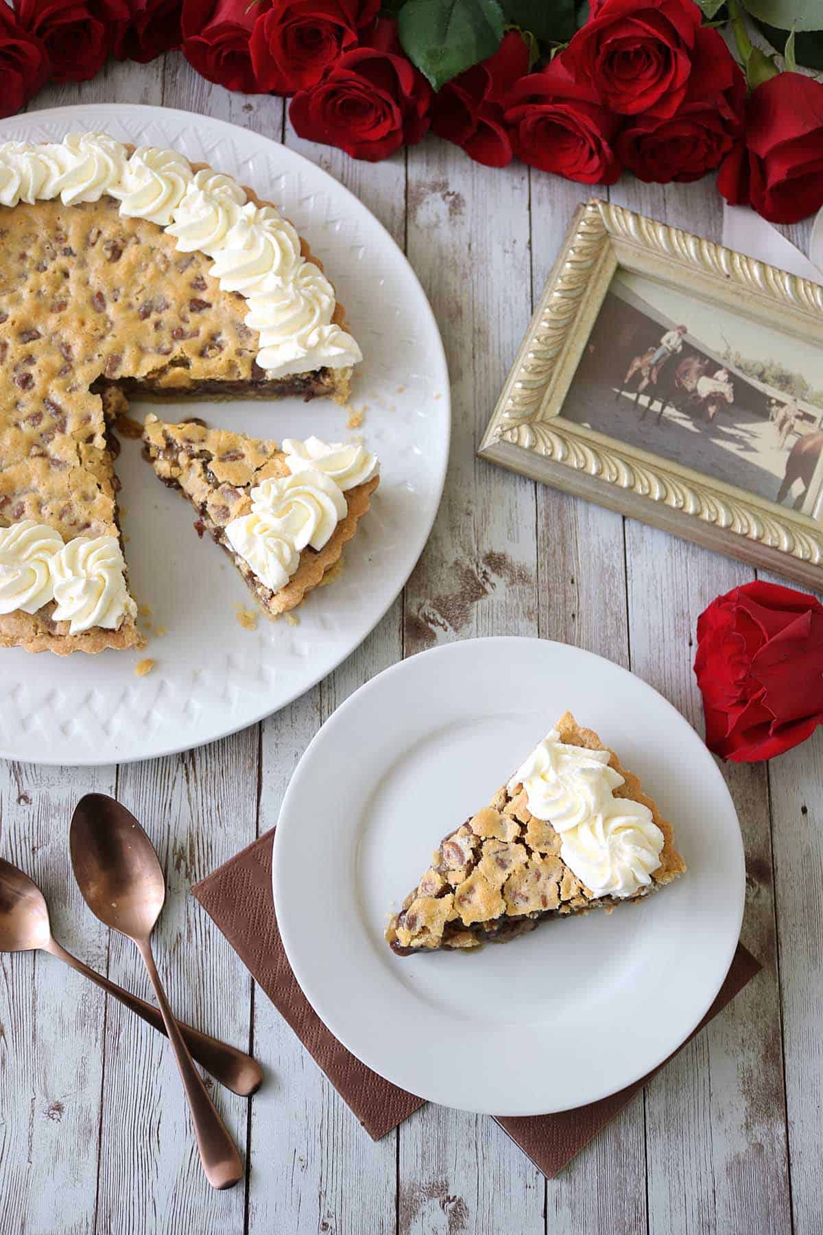 A slice of chocolate chip cookie pie with whipped cream on a white plate, alongside the rest of the pie, two spoons, red roses, and a framed vintage photo on a rustic wooden table.