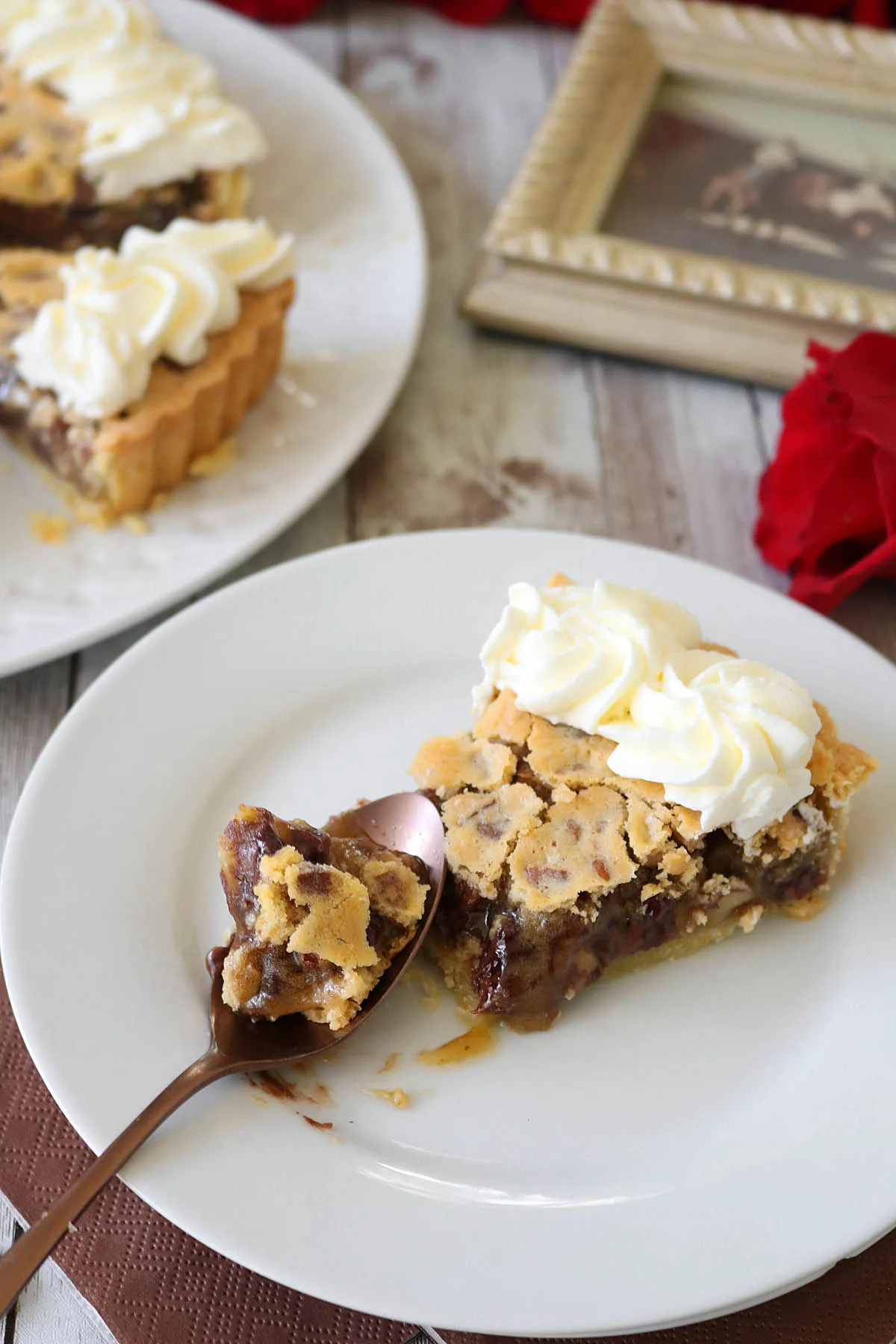 A slice of pie topped with whipped cream sits on a white plate with a spoonful taken out, showing the gooey filling. In the background, more pie is on a platter, next to a red rose and a framed photo.