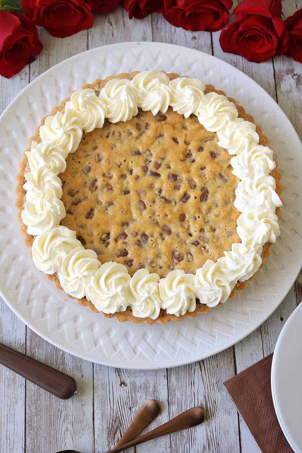 A large chocolate chip cookie cake with piped swirls of white frosting around the edge, displayed on a white plate with red roses in the background on a rustic wooden table.