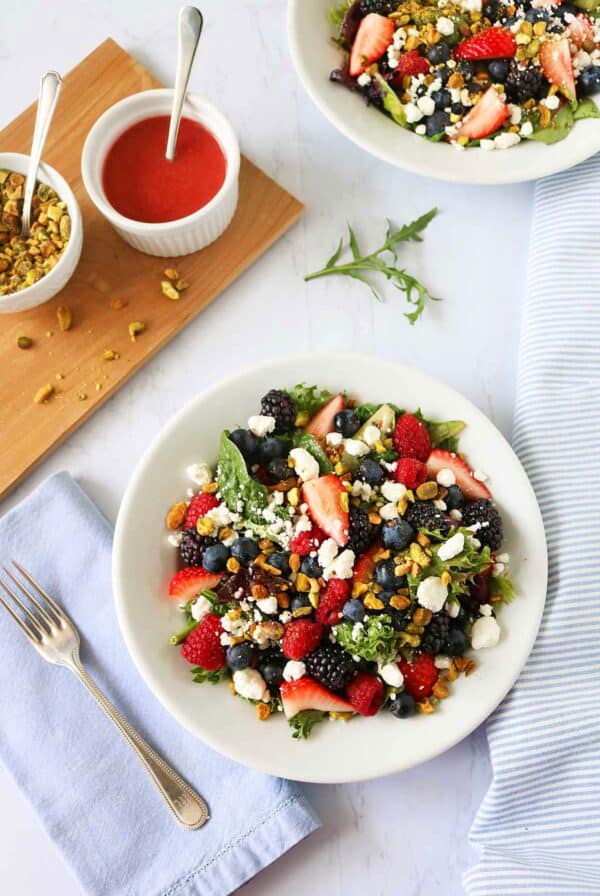 A fresh Berry and Pistachio Salad with mixed greens, blackberries, strawberries, blueberries, goat cheese, and crunchy granola sits on a white plate next to a fork, light blue napkin, extra granola, and a small bowl of red dressing.