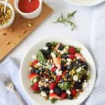 A fresh Berry Goat Cheese and Pistachio Salad with mixed greens, strawberries, blackberries, blueberries, goat cheese, and nuts in a white bowl, next to a fork, blue napkin, and a small dish of red dressing on a white surface.