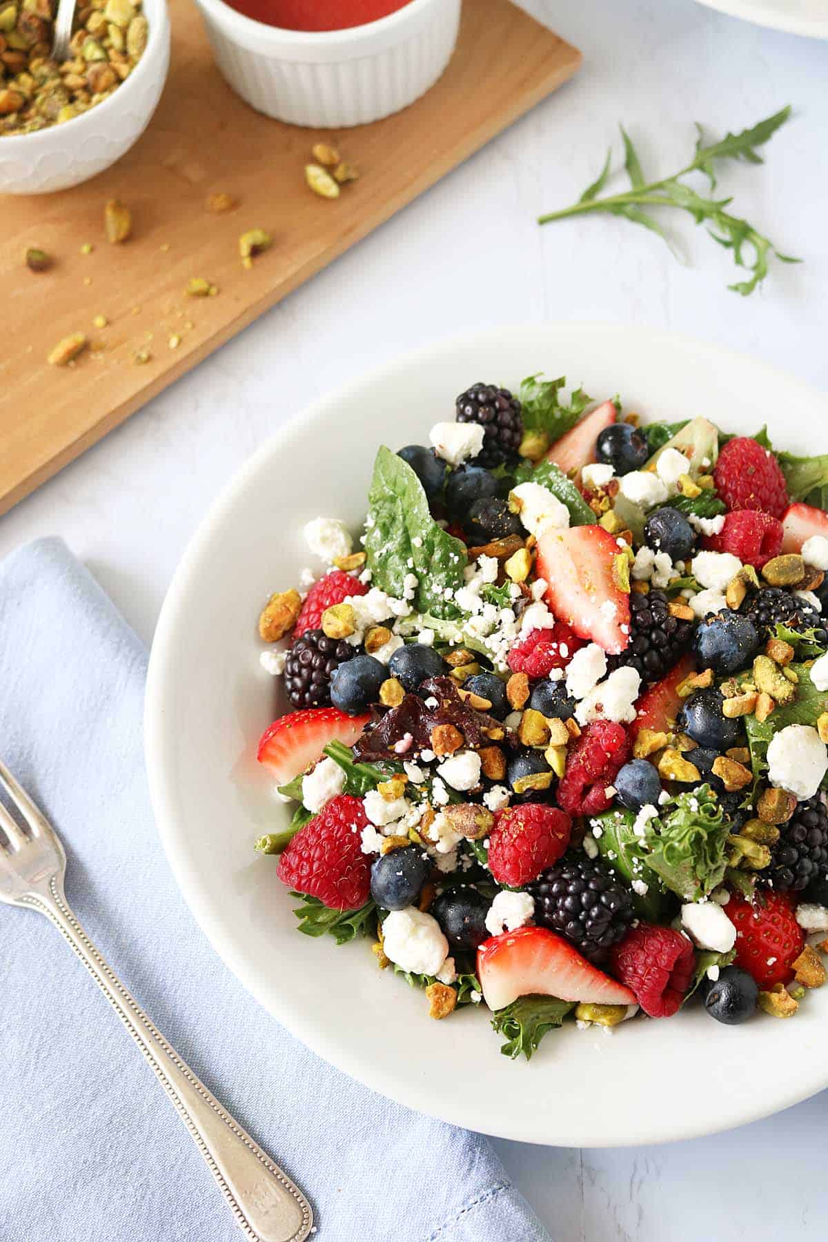 A fresh Berry and Pistachio Salad on a white plate, topped with strawberries, blueberries, blackberries, raspberries, creamy goat cheese, and chopped nuts, placed next to a fork and a light blue napkin.