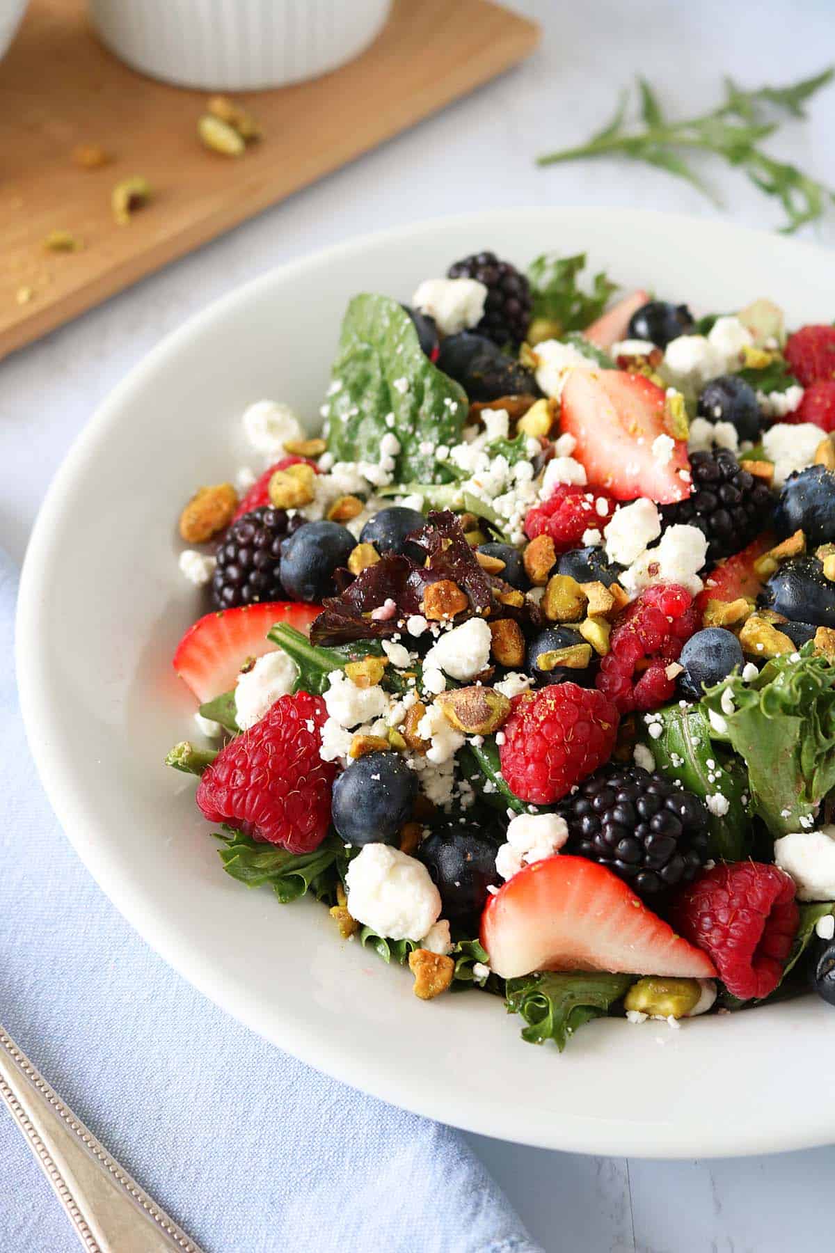 A fresh Berry, Goat Cheese, and Pistachio Salad on a white plate, topped with raspberries, blackberries, blueberries, strawberry slices, leafy greens, and chopped pistachios. A cutting board and herbs are in the background.
