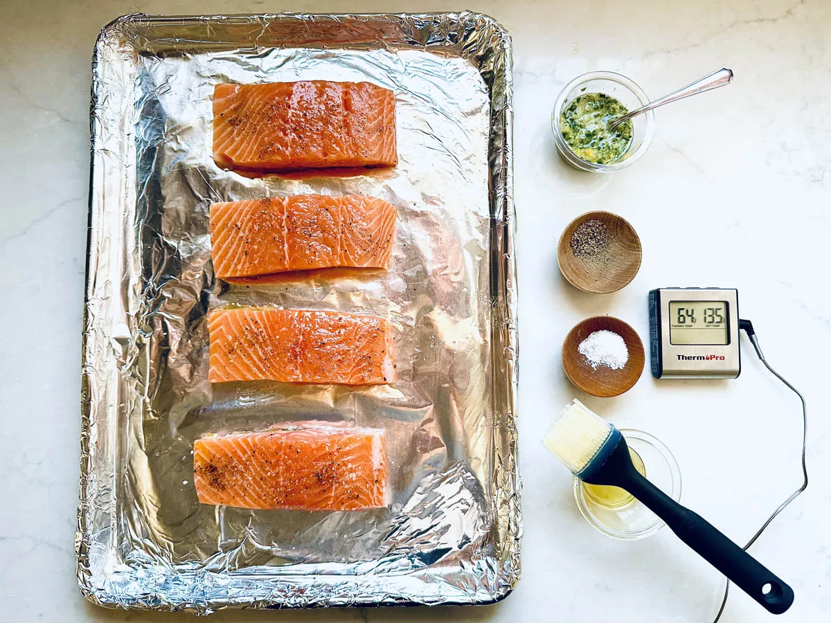 Four raw fillets for Oven-Baked Salmon with Lemon, Fresh Herbs, and Garlic rest on a foil-lined tray, surrounded by bowls of seasoning, melted butter, herb mixture, a basting brush, and a digital thermometer on a white countertop.