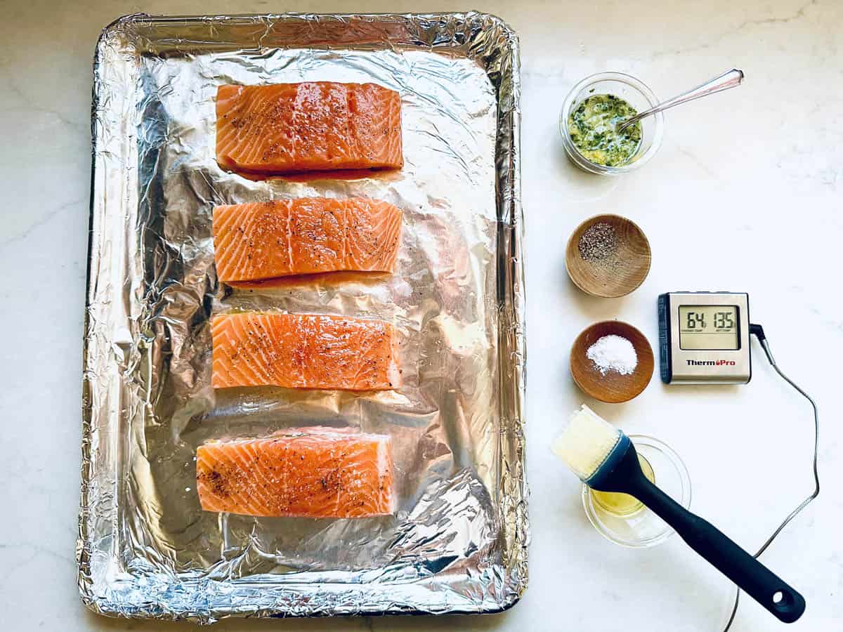 Four raw fillets for Oven-Baked Salmon with Lemon, Fresh Herbs, and Garlic rest on a foil-lined tray, surrounded by bowls of seasoning, melted butter, herb mixture, a basting brush, and a digital thermometer on a white countertop.