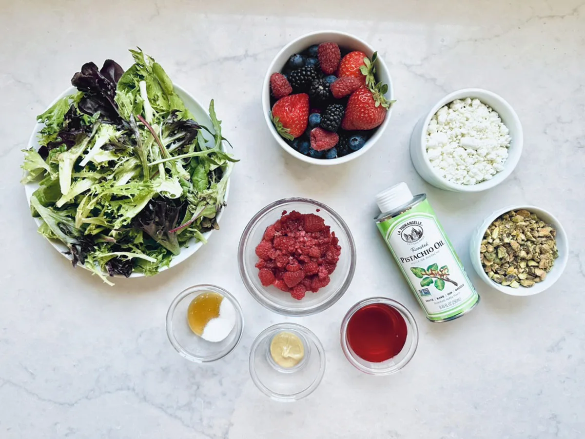A top-down view of a Goat Cheese and Pistachio Salad: mixed greens, fresh berry slices, crumbled goat cheese, chopped pistachios, raspberry vinaigrette components, a small bottle of pistachio oil, plus bowls of honey, salt, and mustard.