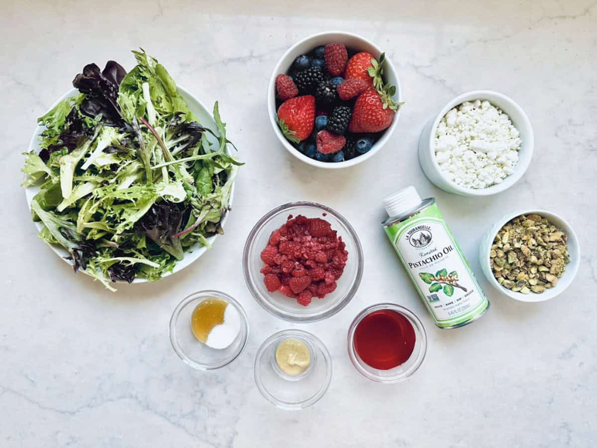 A top-down view of a Goat Cheese and Pistachio Salad: mixed greens, fresh berry slices, crumbled goat cheese, chopped pistachios, raspberry vinaigrette components, a small bottle of pistachio oil, plus bowls of honey, salt, and mustard.