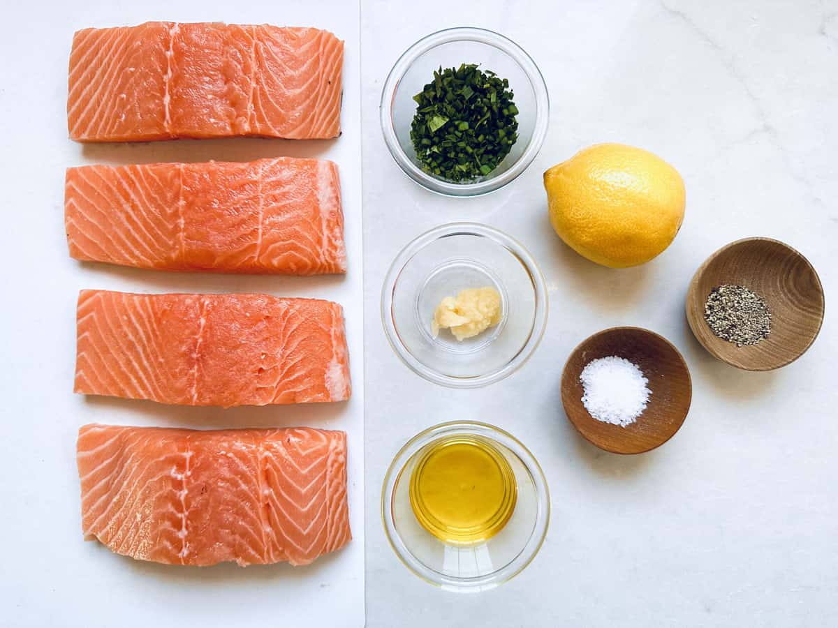 Oven-Baked Salmon with Lemon and fresh herbs: Four salmon fillets, a bowl of chopped herbs, a lemon, minced garlic, ground pepper, salt, and olive oil arranged neatly on a white surface.