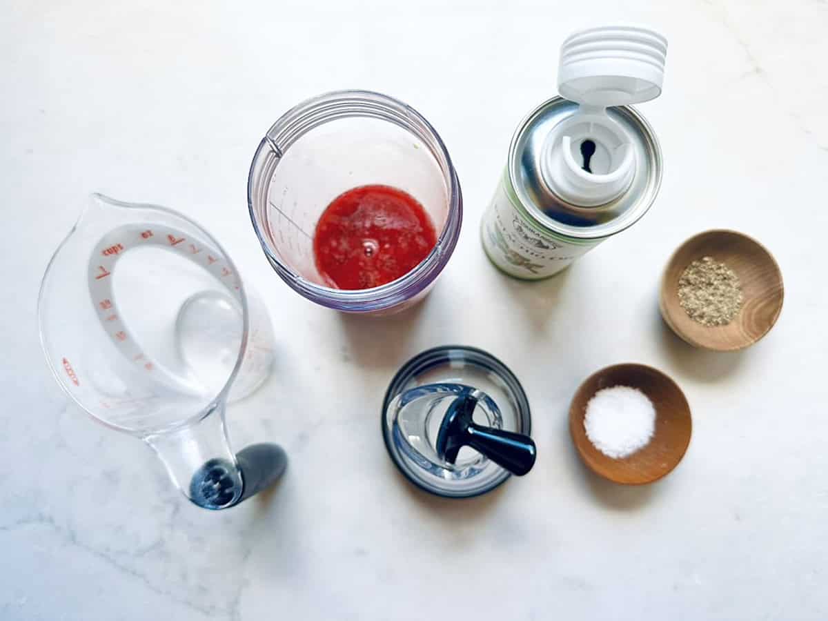 Top-down view of a measuring cup with water, a blender cup with red berry puree, an oil spray can, and three small bowls containing black pepper, salt, and white seasoning—perfect for making a Goat Cheese and Pistachio Salad.