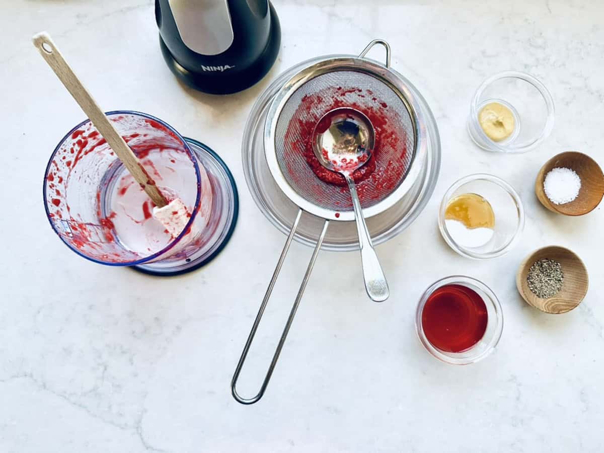 A blender cup with red berry puree, a strainer with red residue, spoon, and small bowls with mustard, honey, red vinegar, salt, and pepper are arranged on a white countertop—perfect for dressing a Goat Cheese and Pistachio Salad.