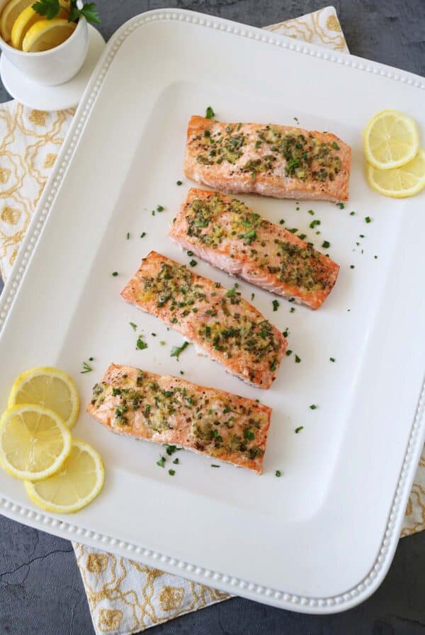 Four oven-baked salmon fillets with lemon and fresh herbs are arranged on a white platter. Lemon slices and chopped parsley add brightness, with a patterned napkin and a small bowl of lemons nearby.