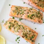Three oven-baked salmon fillets topped with garlic and fresh herbs are arranged on a white plate, garnished with chives. Two lemon slices rest beside the salmon, enhancing this Oven-Baked Salmon with Lemon, Garlic, and Fresh Herbs dish.
