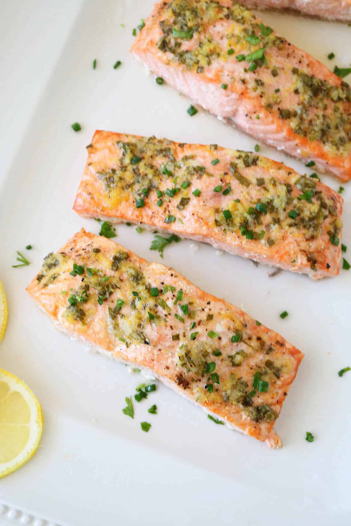 Three oven-baked salmon fillets topped with garlic and fresh herbs are arranged on a white plate, garnished with chives. Two lemon slices rest beside the salmon, enhancing this Oven-Baked Salmon with Lemon, Garlic, and Fresh Herbs dish.