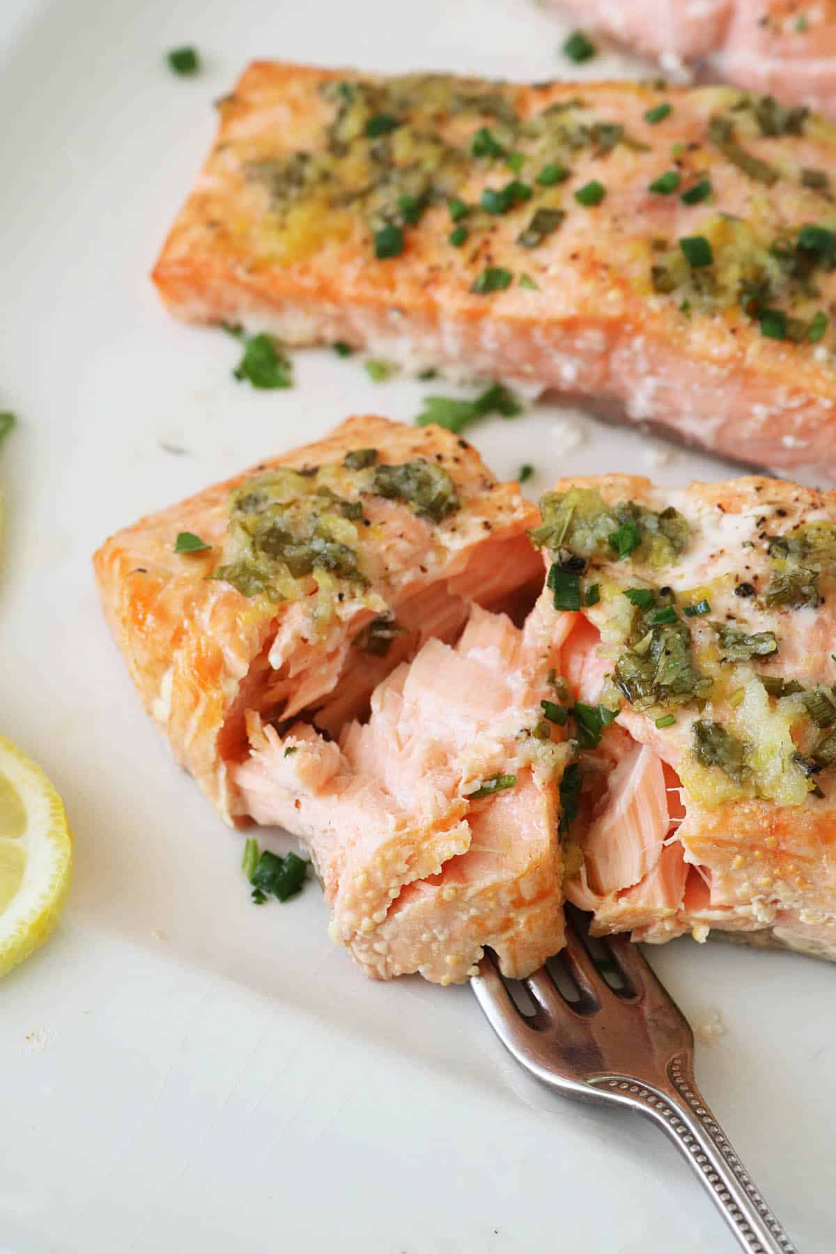 A close-up of oven-baked salmon fillets garnished with garlic and fresh herbs, with a fork flaking a piece. Slices of lemon are visible on the side, and the salmon appears moist and tender on a white plate.