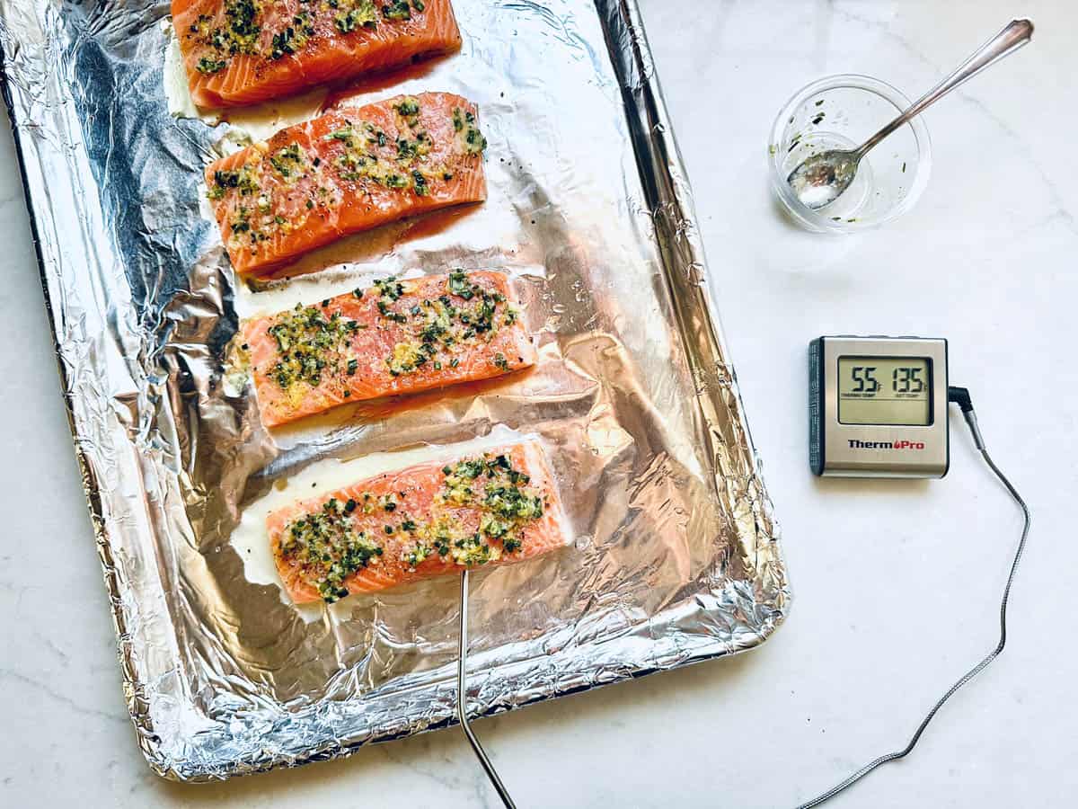Four raw salmon fillets topped with fresh herbs and garlic on a foil-lined baking sheet, with a meat thermometer showing 135°F, and a small bowl of herb mixture nearby—a perfect start for Oven-Baked Salmon with Lemon.