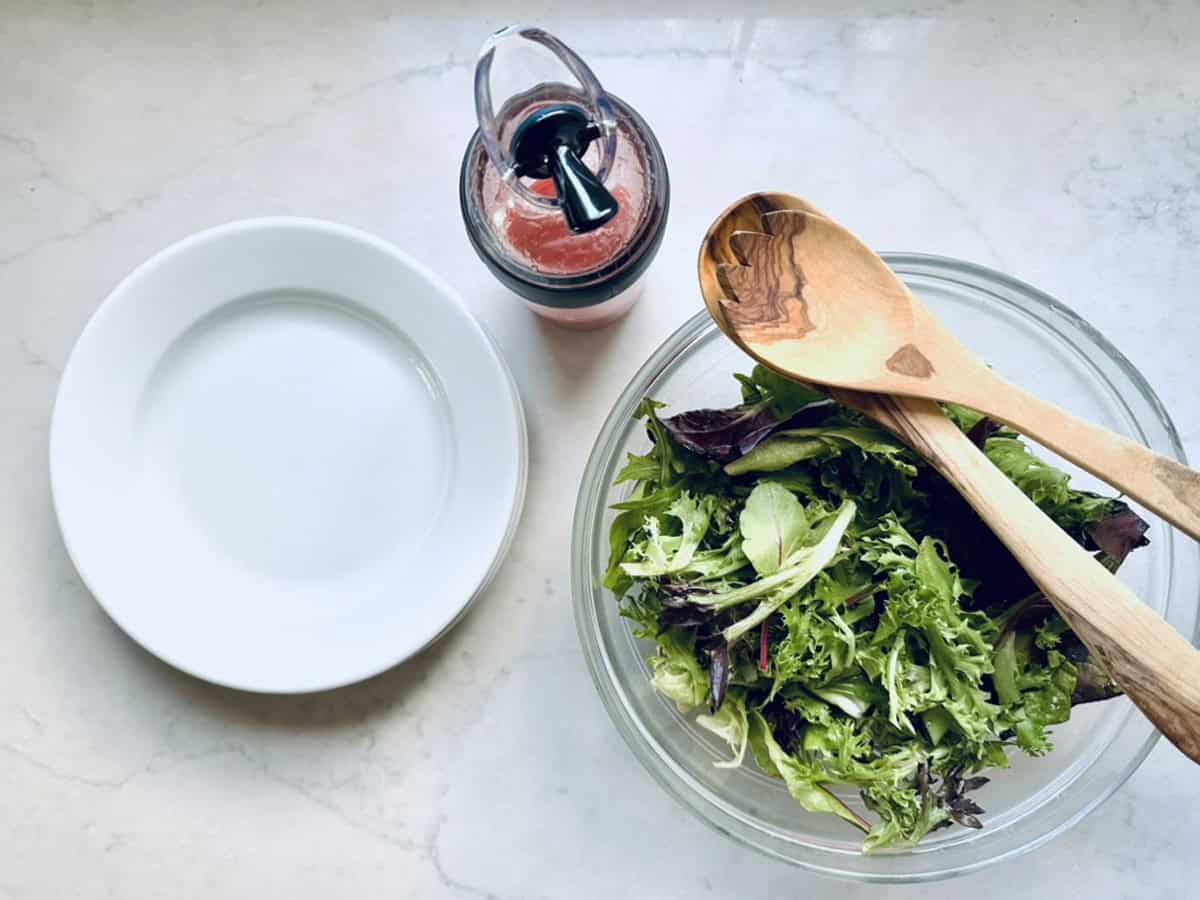 A clear glass bowl of Berry, Goat Cheese, and Pistachio Salad with wooden serving utensils sits beside a white empty plate and a bottle of pink salad dressing on a light countertop.