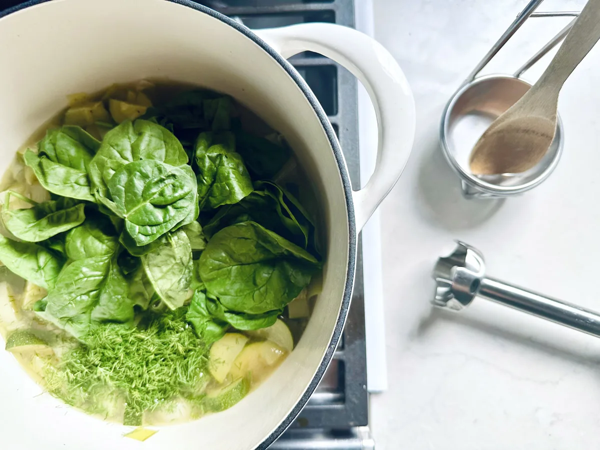 A pot on a stove contains fresh spinach, chopped vegetables, and herbs&mdash;ingredients for a Zucchini Soup with Dill and Greek Yogurt. Nearby are a wooden spoon in a metal holder and an immersion blender, suggesting comforting soup preparation.