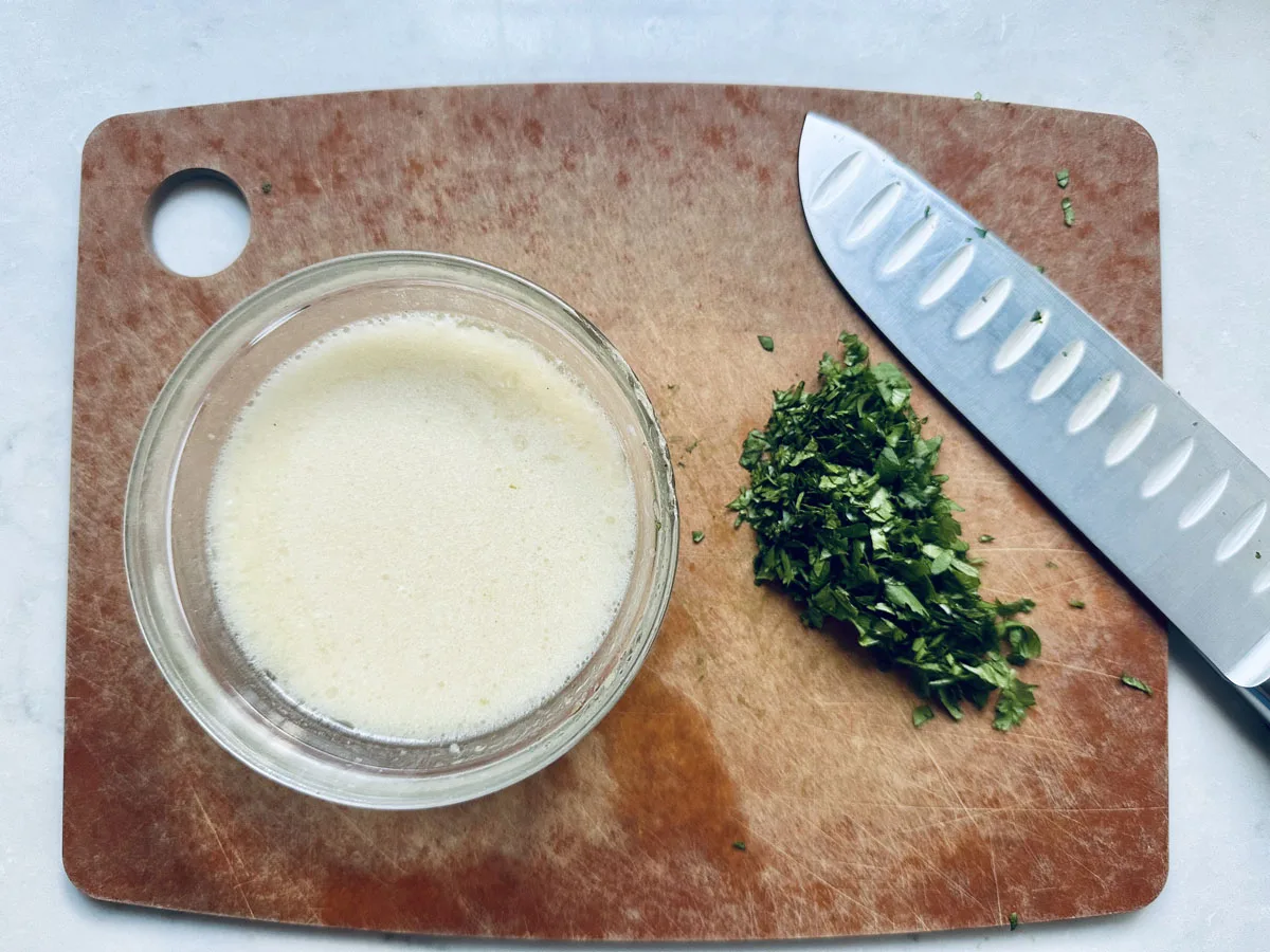 A glass bowl of creamy liquid, chopped herbs, and a knife are on a wooden cutting board with a hole in the top left corner, set on a light countertop.