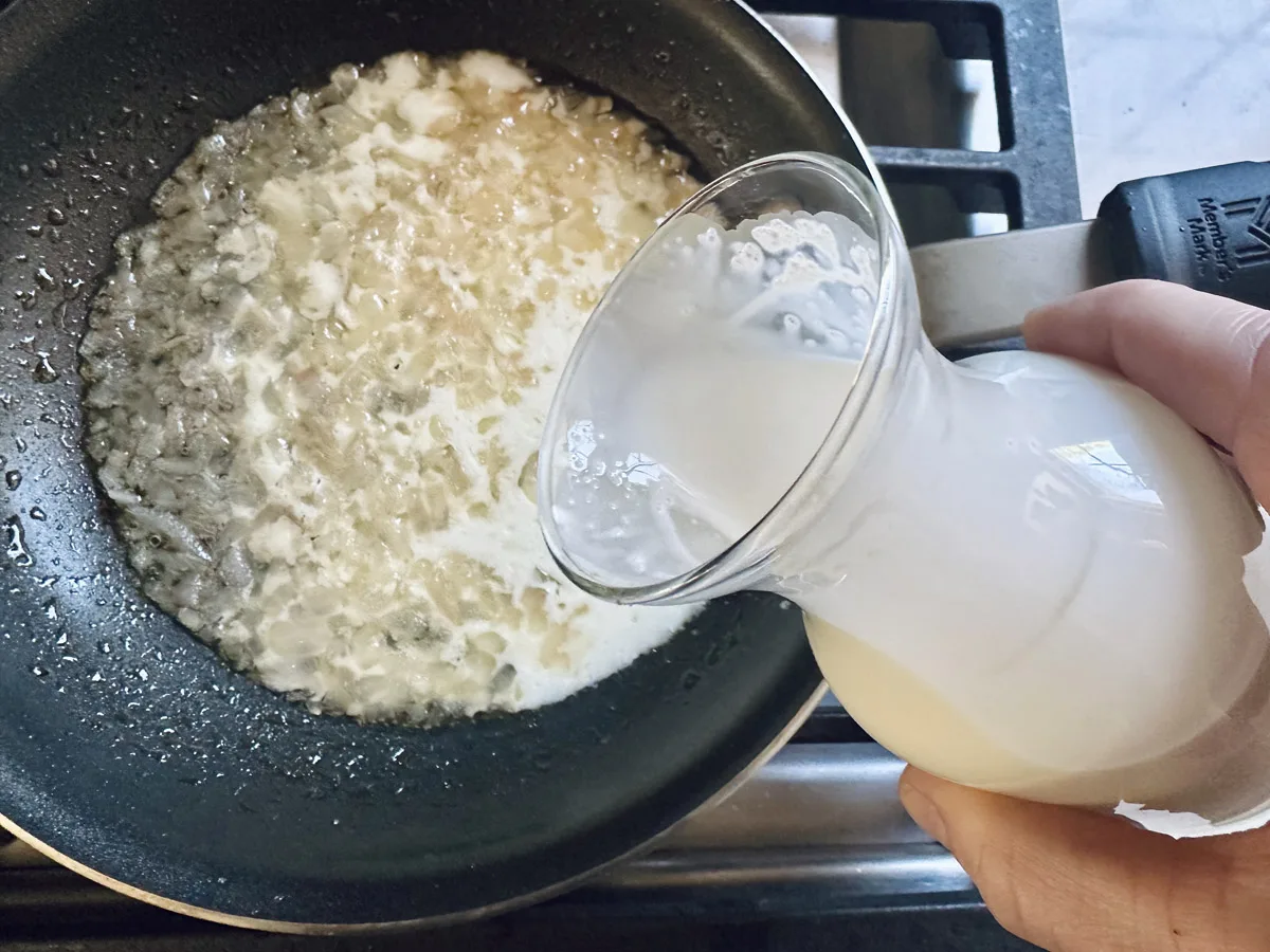 A hand pours cream from a glass pitcher into a frying pan with saut&eacute;ed onions on a stovetop.