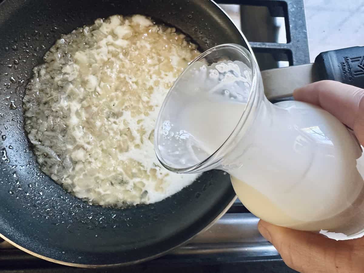 A hand pours cream from a glass pitcher into a frying pan with sautéed onions on a stovetop.