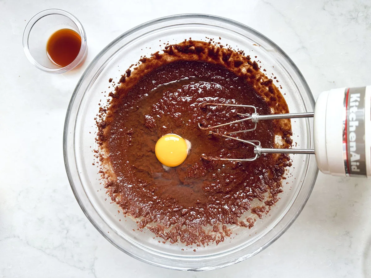 A glass bowl with Chocolate Chipotle Brownies batter and a raw egg in the center, being mixed with an electric hand mixer; a small cup of vanilla extract sits beside the bowl on a white countertop.