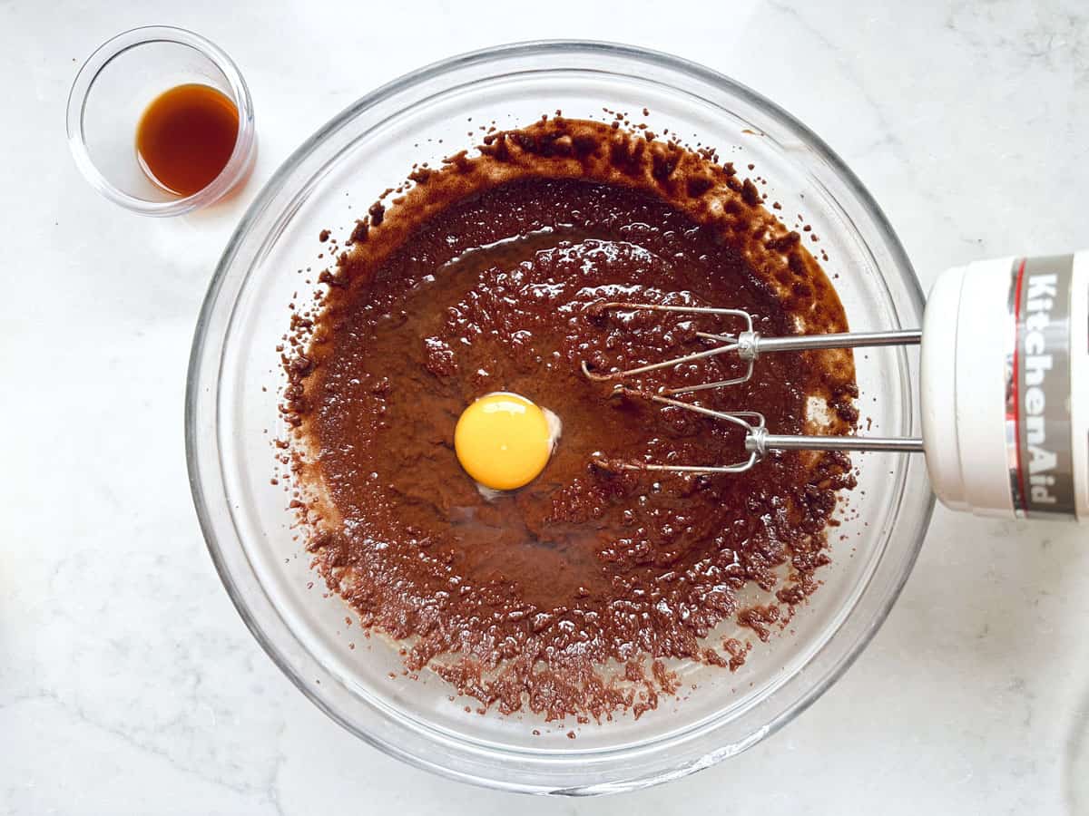 A glass bowl with Chocolate Chipotle Brownies batter and a raw egg in the center, being mixed with an electric hand mixer; a small cup of vanilla extract sits beside the bowl on a white countertop.