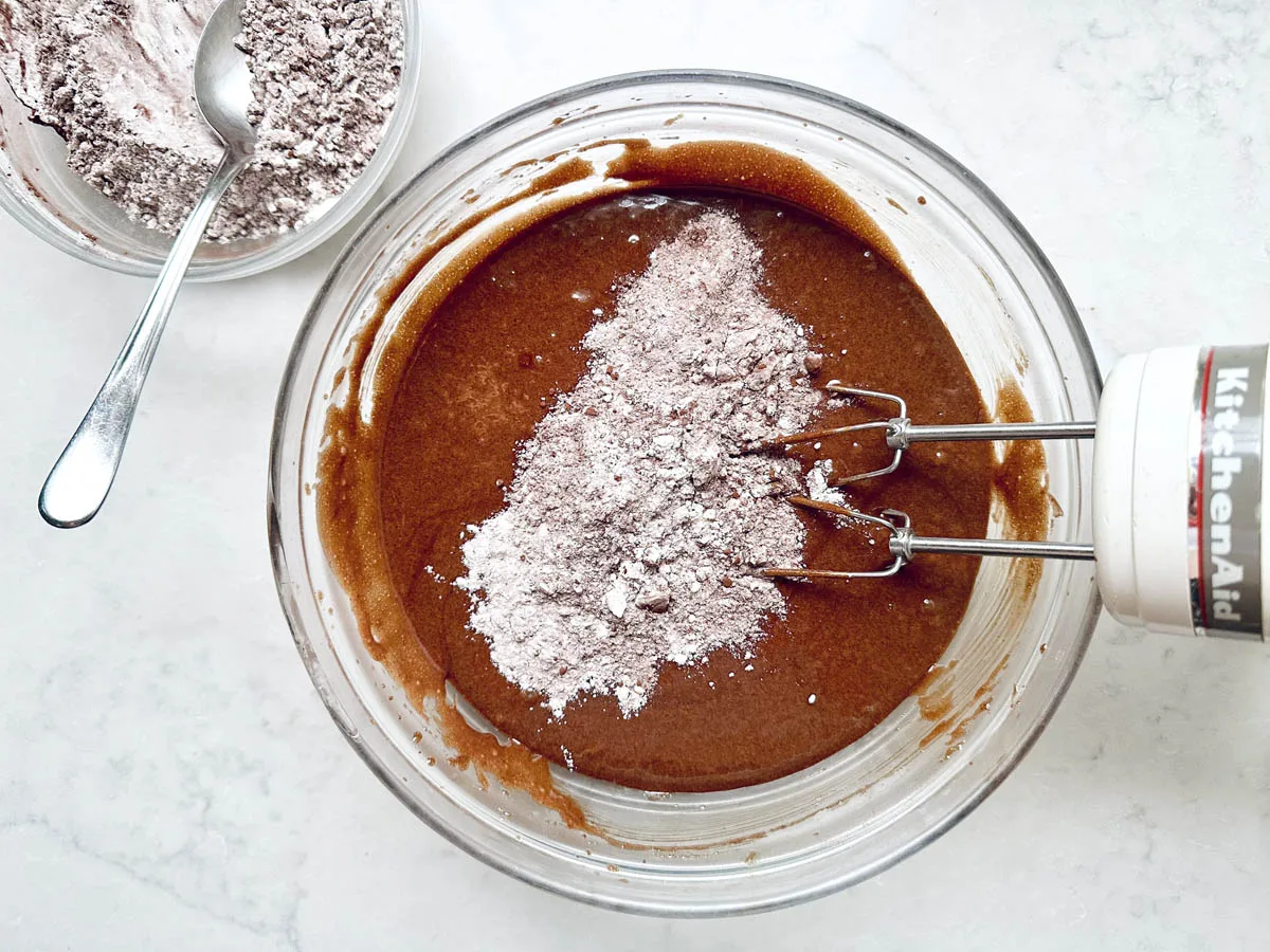 A glass bowl filled with Chocolate Chipotle Brownies batter is being mixed with a hand mixer, topped with dry cocoa powder. A smaller bowl of more dry ingredients and a spoon rests nearby on the white countertop.