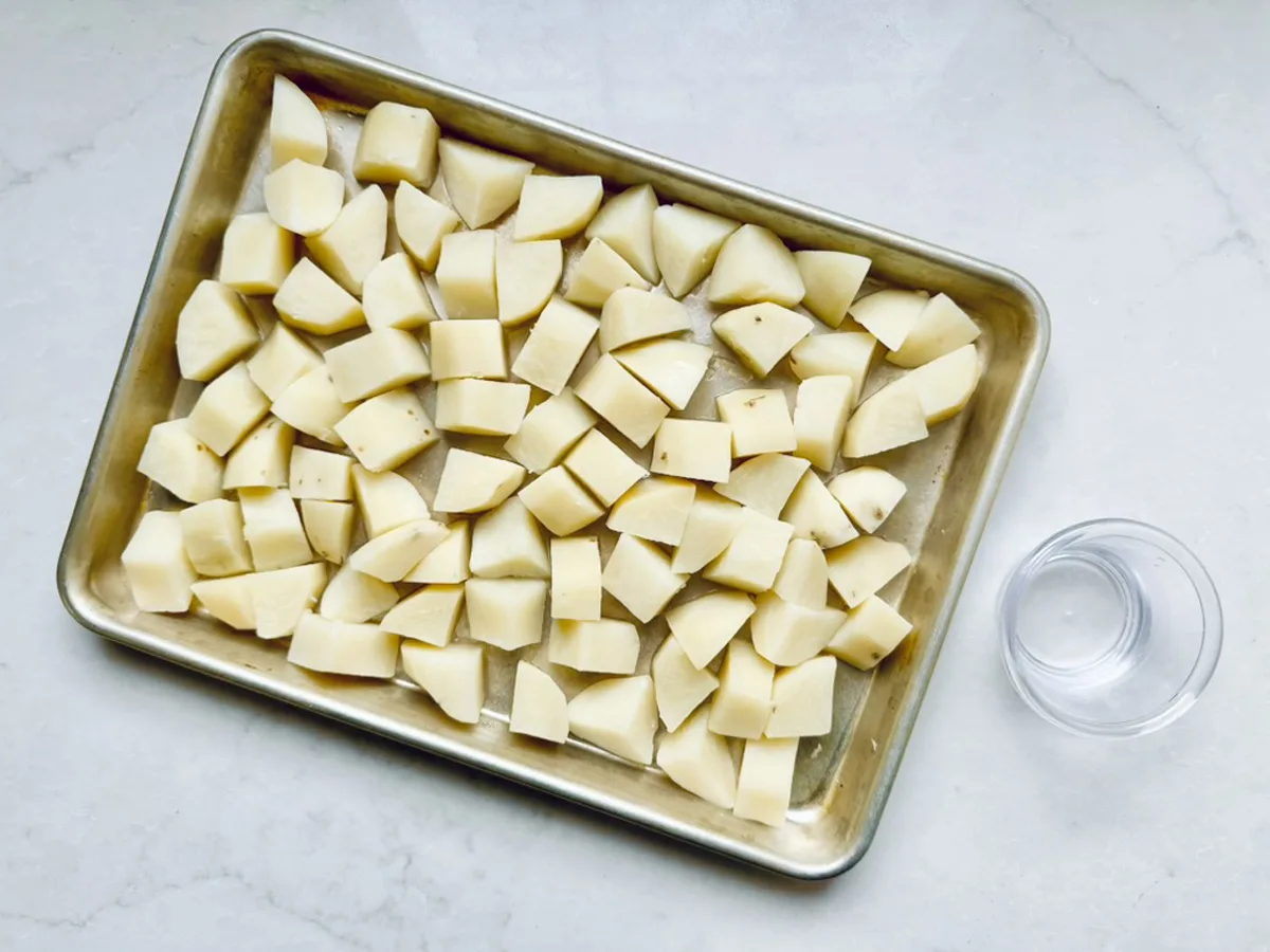 A baking tray filled with evenly spread, raw potato chunks&mdash;perfect for All-American Russet Potato Salad&mdash;sits on a light-colored countertop beside a small clear glass bowl of water.