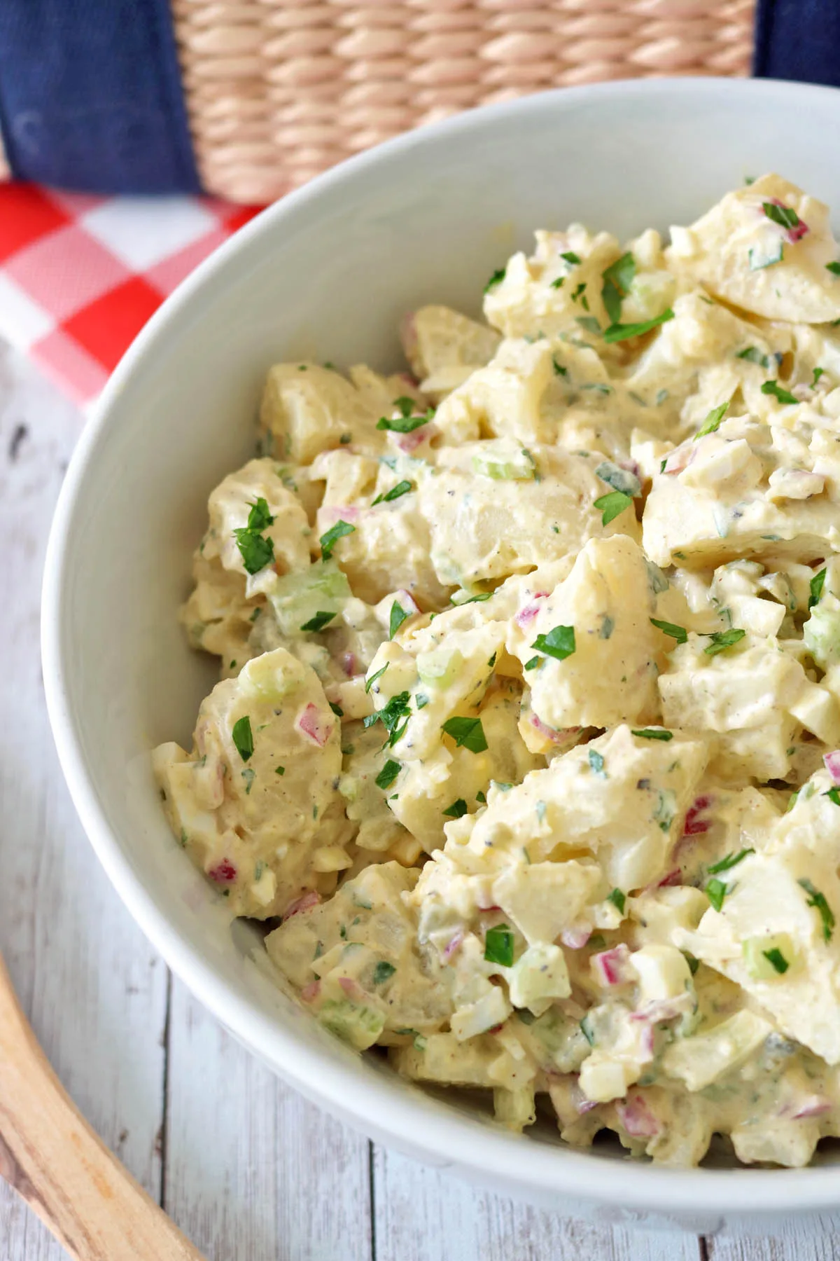 A close-up of a bowl of All-American Russet Potato Salad, creamy and topped with chopped parsley, diced red onions, and celery, set on a white wooden table with a red and white checkered napkin nearby.