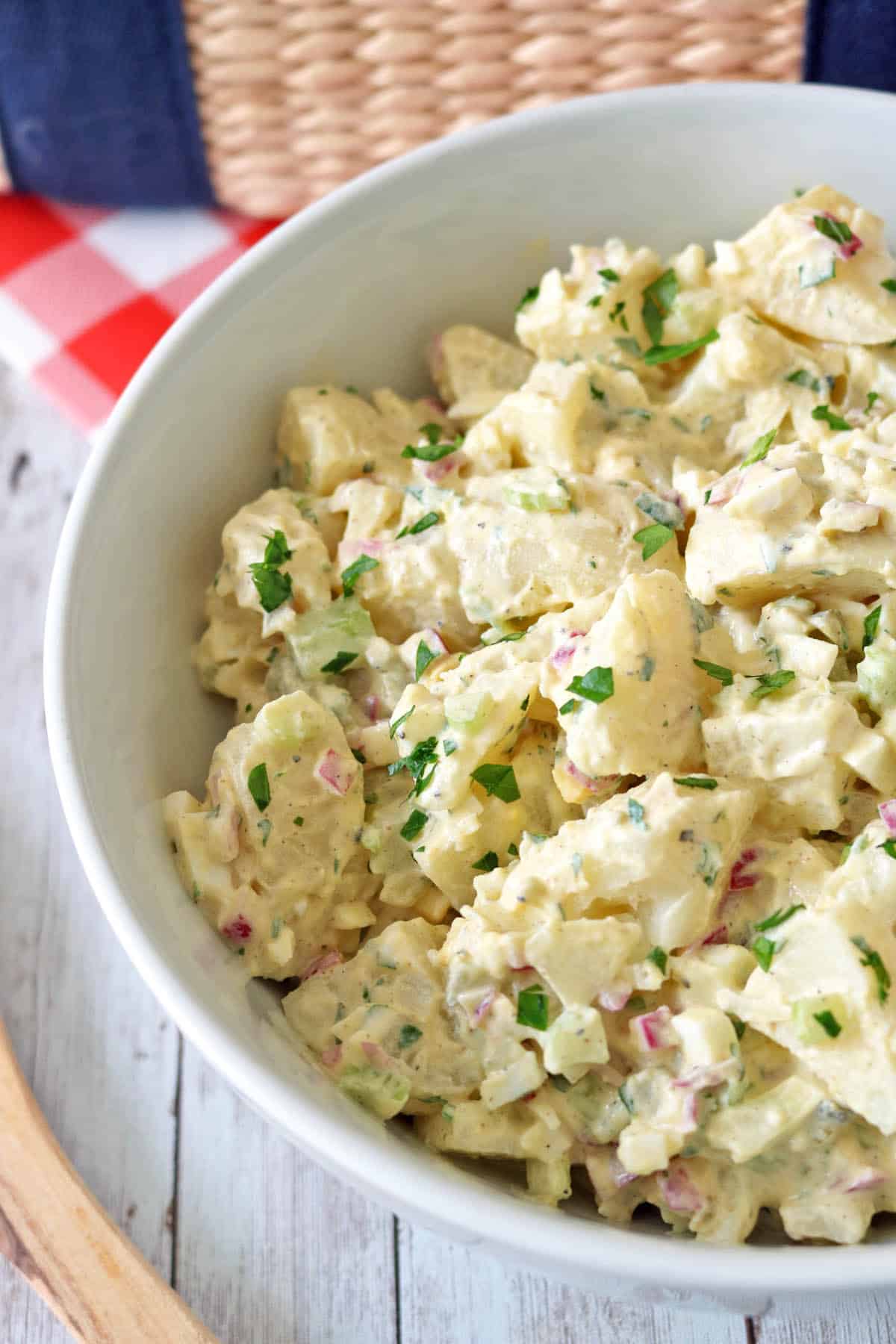 A close-up of a bowl of All-American Russet Potato Salad, creamy and topped with chopped parsley, diced red onions, and celery, set on a white wooden table with a red and white checkered napkin nearby.