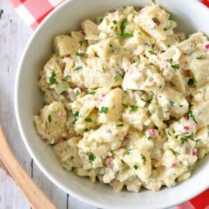 A white bowl of All-American Russet Potato Salad with chopped red onions, celery, and parsley sits on a rustic white wooden table with a red-checked napkin and a woven basket in the background.