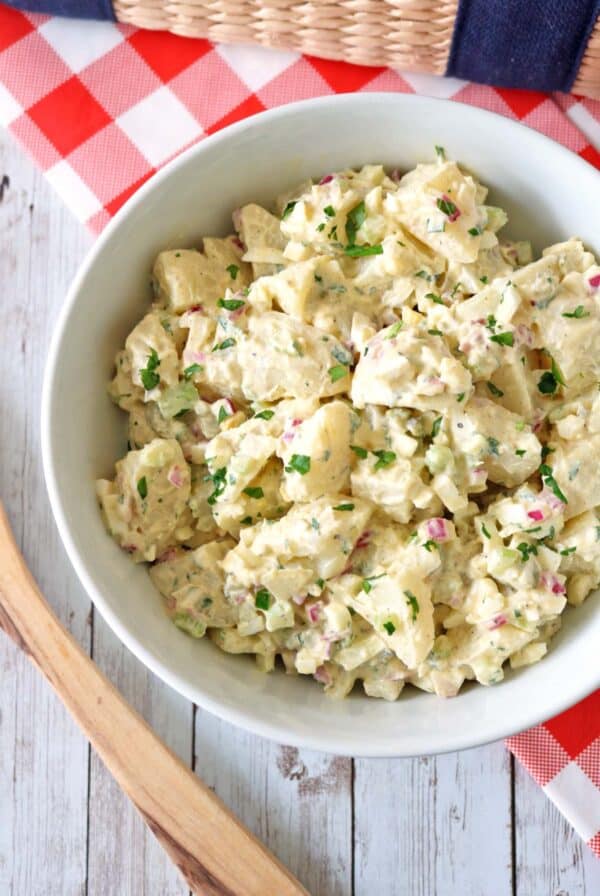 A white bowl of All-American Russet Potato Salad with chopped red onions, celery, and parsley sits on a rustic white wooden table with a red-checked napkin and a woven basket in the background.