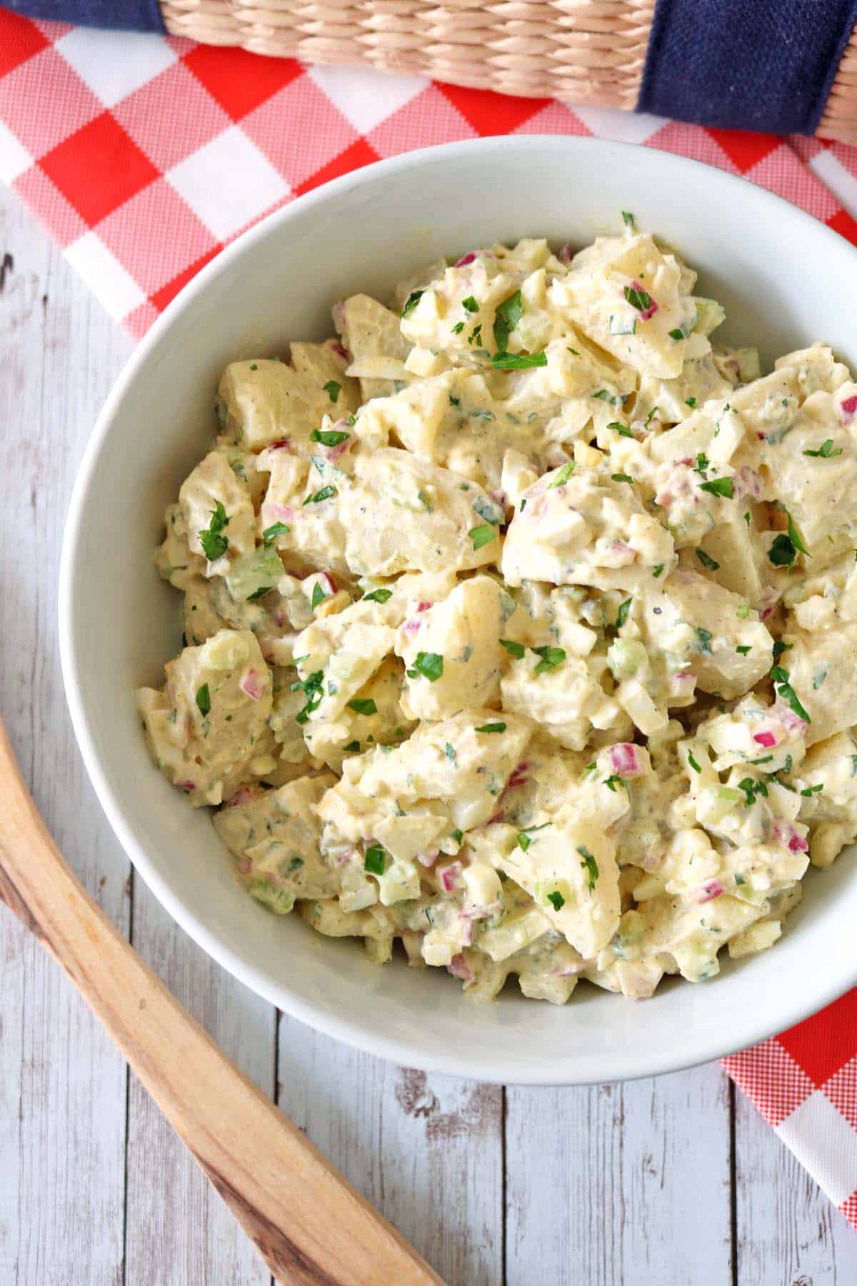 A white bowl of All-American Russet Potato Salad with chopped red onions, celery, and parsley sits on a rustic white wooden table with a red-checked napkin and a woven basket in the background.