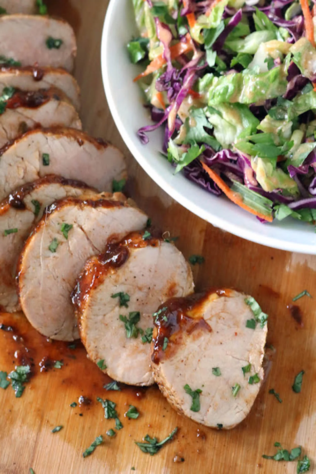 Sliced pork tenderloin with a glossy sauce, garnished with herbs, is served on a wooden board next to a bowl of colorful coleslaw with cabbage, carrots, and greens.
