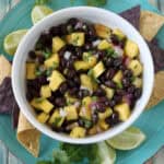 A bowl of black bean and mango salsa with chopped onions and cilantro, served on a blue plate with tortilla chips, lime wedges, and cilantro sprigs on a rustic wooden table.