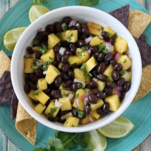 A bowl of black bean and mango salsa with chopped onions and cilantro, served on a blue plate with tortilla chips, lime wedges, and cilantro sprigs on a rustic wooden table.
